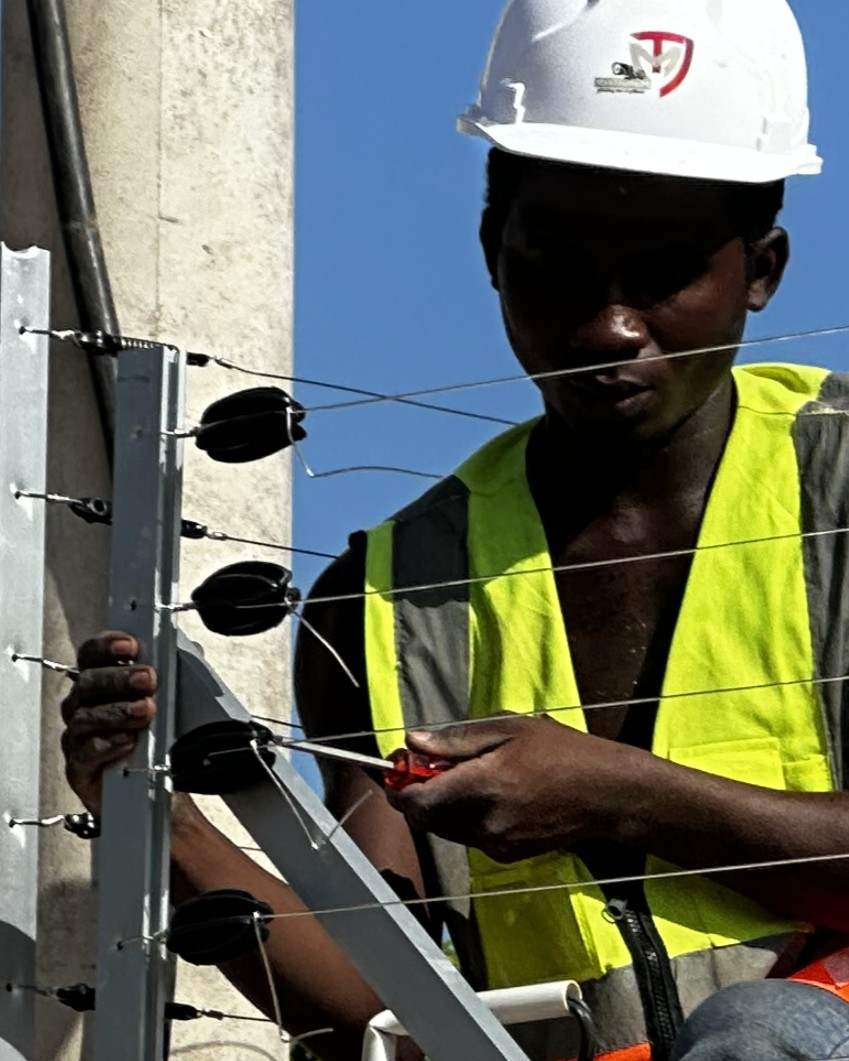 Fundi aliyevaa kofia ngumu na fulana inayoakisi mwanga akiunganisha nyaya za uzio wa umeme
