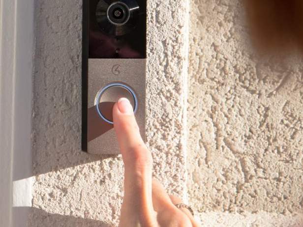 A finger pressing a video doorbell mounted beside a door